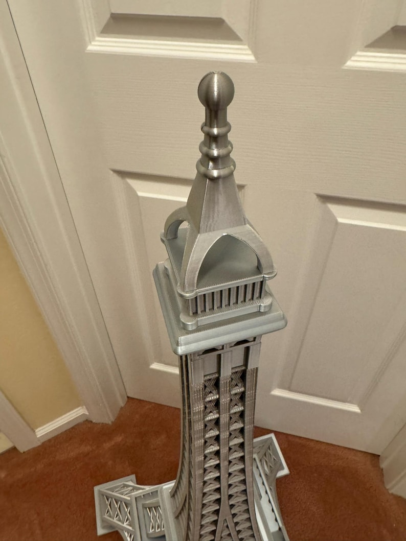 May include: A silver-colored model of the Eiffel Tower. The tower is intricately detailed, with a lattice-like structure and architectural features. The model is standing on a brown carpet in front of a white door.