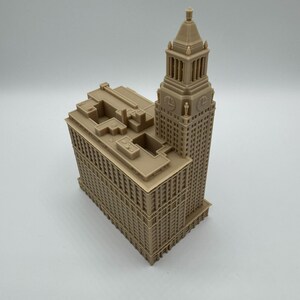 May include: A tan-colored architectural model of a tall building with a tower. The model has detailed windows and architectural features. The building's design includes a tower with a spire, and a rectangular base.