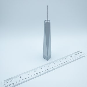 May include: A silver model of the One World Trade Center building, approximately 6 inches tall, with a ruler for scale.