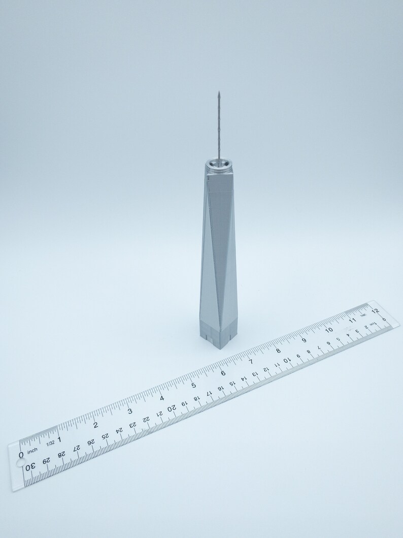 May include: A silver model of the One World Trade Center building, approximately 6 inches tall, with a ruler for scale.
