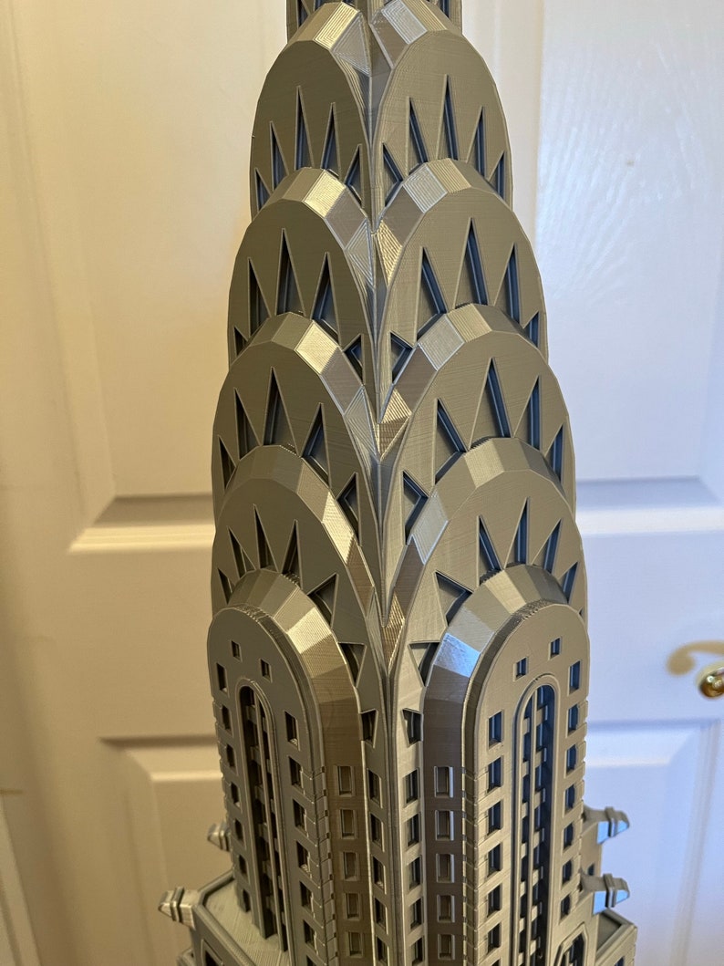 May include: A 3D printed model of the Chrysler Building in a silver metallic finish. The model features the building's iconic Art Deco design, including the pointed spire and the stepped setbacks.