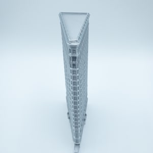 Puede incluir: Un modelo 3D impreso en gris del Flatiron Building en la ciudad de Nueva York. El modelo es un prisma triangular con un techo plano y muchas ventanas.