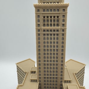 May include: A beige architectural model of a tall building with multiple stories and rectangular windows. The building has a central tower and several lower wings, all in a similar color and style. The model is likely a replica of a famous landmark.