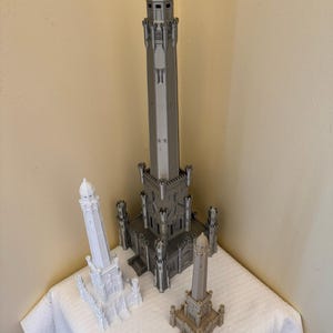 May include: Three miniature architectural models in varying sizes and colors. The largest is gray, the medium is white, and the smallest is tan. A ruler is placed in front of the models, indicating their scale. The models are displayed on a white surface.