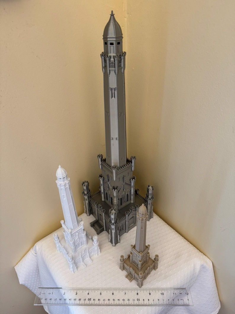 May include: Three miniature architectural models in varying sizes and colors. The largest is gray, the medium is white, and the smallest is tan. A ruler is placed in front of the models, indicating their scale. The models are displayed on a white surface.