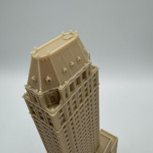 Może przedstawiać: Beżowy, drukowany w 3D model wysokiego budynku ze szczeg&oacute;łową fasadą i charakterystycznym dachem. Budynek posiada wiele okien i detali architektonicznych, prezentujących klasyczny projekt wieżowca.