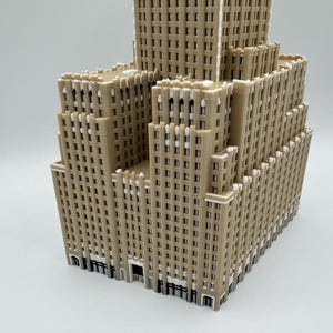 May include: A tan-coloured, multi-storey building model with a detailed facade. The model features numerous windows and architectural details, with a tiered design. The building has a light brown colour with white accents.