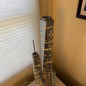 May include: A set of illuminated model skyscrapers, featuring a large central building and several smaller replicas. The buildings are gray with lit-up window details, creating a warm glow. The models are placed on a white surface, with a ruler and power strip visible.