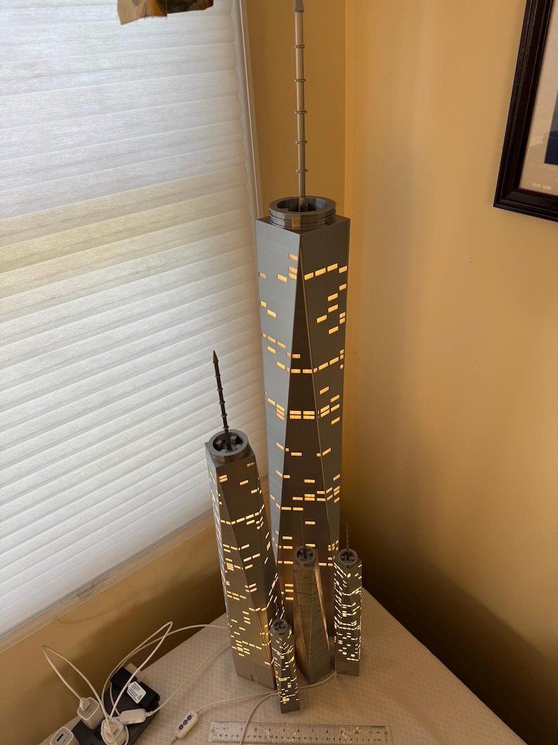 May include: A set of illuminated model skyscrapers, featuring a large central building and several smaller replicas. The buildings are gray with lit-up window details, creating a warm glow. The models are placed on a white surface, with a ruler and power strip visible.