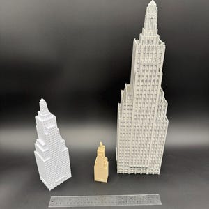 May include: Three miniature skyscraper models in white and tan, with a ruler for scale. The tallest building is a light gray color, and the smallest is tan. The buildings have detailed architectural features.