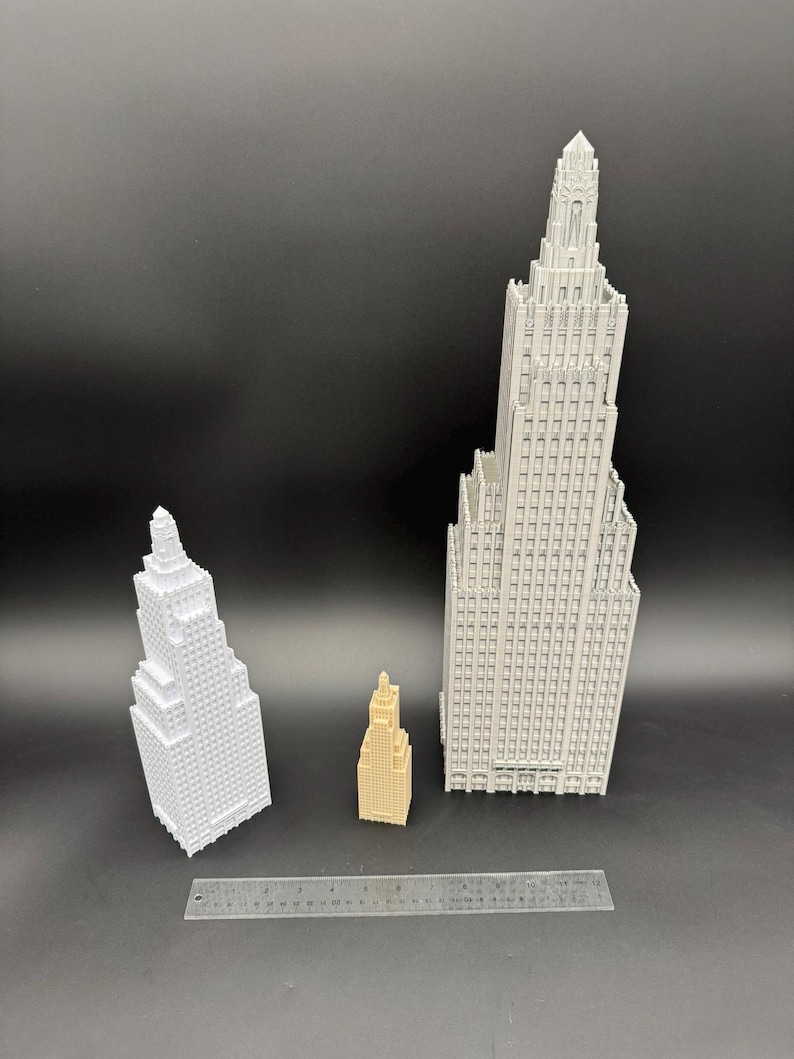May include: Three miniature skyscraper models in white and tan, with a ruler for scale. The tallest building is a light gray color, and the smallest is tan. The buildings have detailed architectural features.