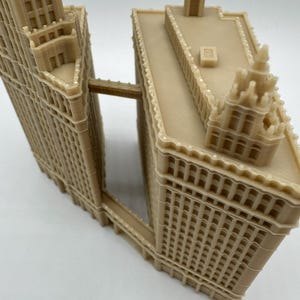 May include: A beige 3D-printed model of a building complex, featuring two tall, rectangular structures connected by a bridge. The model has detailed architectural features, including windows, decorative trim, and rooftop elements. The overall design is intricate and precise.