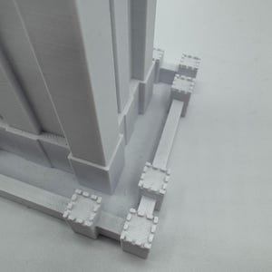 May include: White 3D printed model of a tall building with a square base. The base has a decorative border with small square turrets.