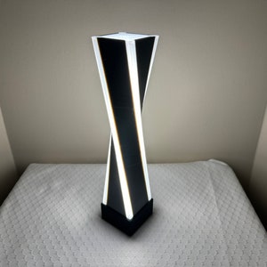 May include: A white and black modern table lamp with a twisted design. The lamp is turned on and illuminating the room.