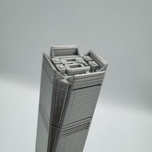 May include: A gray, 3D-printed model of a tall skyscraper. The building has a rectangular shape with detailed horizontal lines and architectural features at the top. The model is set against a plain gray background.