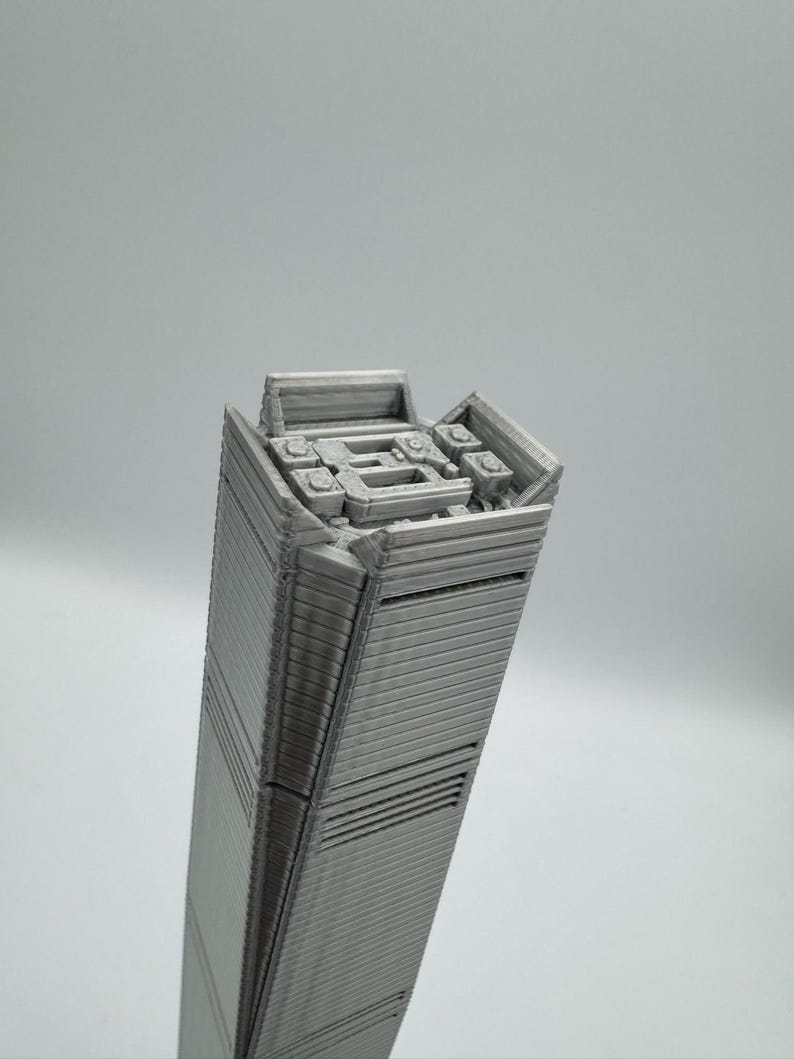 May include: A gray, 3D-printed model of a tall skyscraper. The building has a rectangular shape with detailed horizontal lines and architectural features at the top. The model is set against a plain gray background.