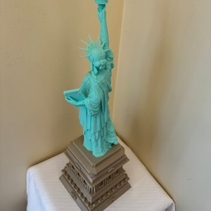 May include: A teal green 3D printed model of the Statue of Liberty standing on a brown base.