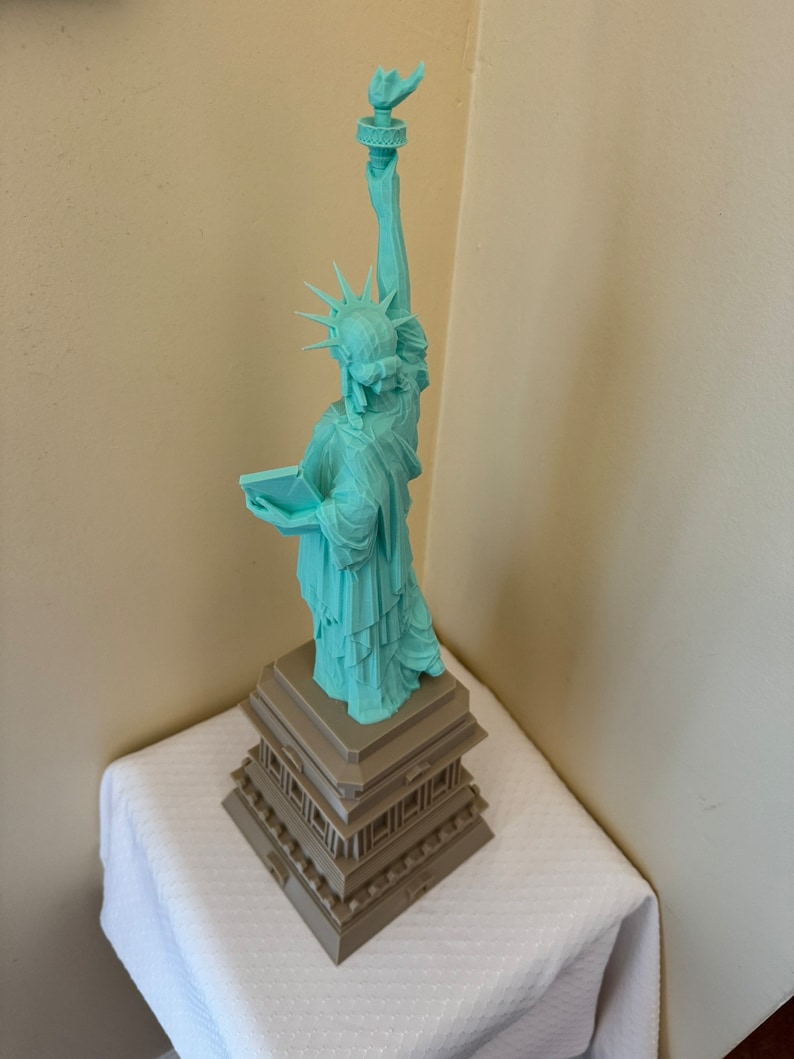 May include: A teal green 3D printed model of the Statue of Liberty standing on a brown base.