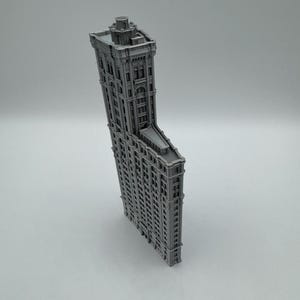 May include: A grey miniature model of the Flatiron Building. The model has detailed architectural features, including windows and cornices, and a distinctive triangular shape. The building is a classic example of early skyscraper design.