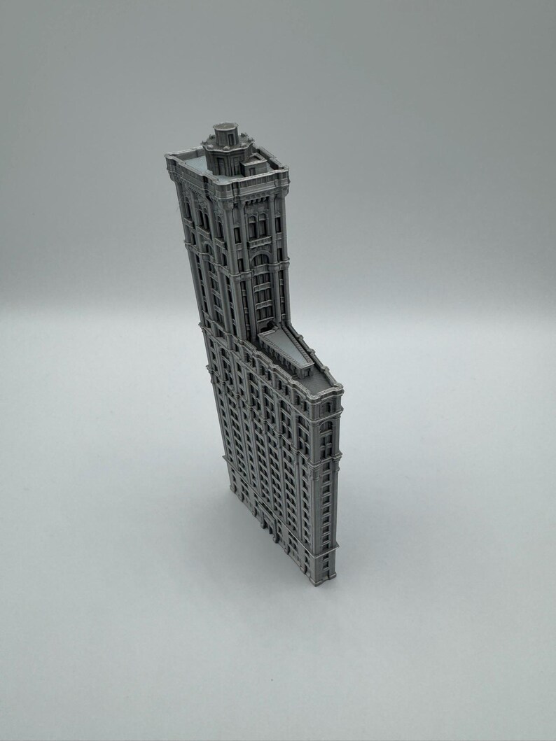 May include: A grey miniature model of the Flatiron Building. The model has detailed architectural features, including windows and cornices, and a distinctive triangular shape. The building is a classic example of early skyscraper design.