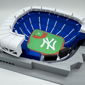 Peut inclure: Un modèle imprimé en 3D d'un stade de baseball avec une structure de toit blanche, des sections de sièges bleu foncé et un terrain vert avec un champ intérieur marron. Le centre du terrain affiche un grand logo NY blanc sur une base grise.