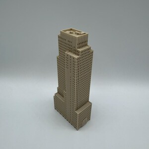 May include: A beige-colored miniature skyscraper model. The building has a rectangular base and a tiered design, with numerous small windows across its facade. The model is set against a plain, light background.
