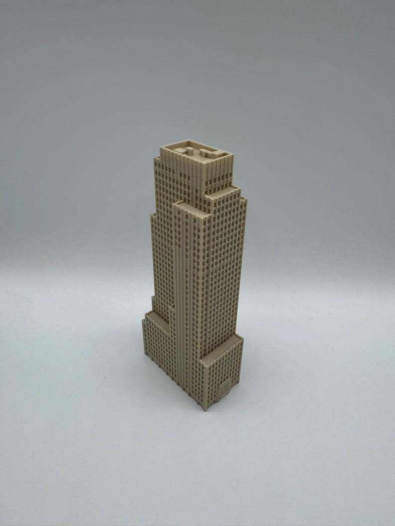 May include: A beige-colored miniature skyscraper model. The building has a rectangular base and a tiered design, with numerous small windows across its facade. The model is set against a plain, light background.