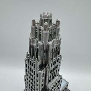 May include: A detailed, gray model of a skyscraper, showcasing intricate architectural details. The building features numerous windows and a tiered design, with a complex crown. The model is likely 3D-printed, with visible layer lines. A miniature replica of a famous building.
