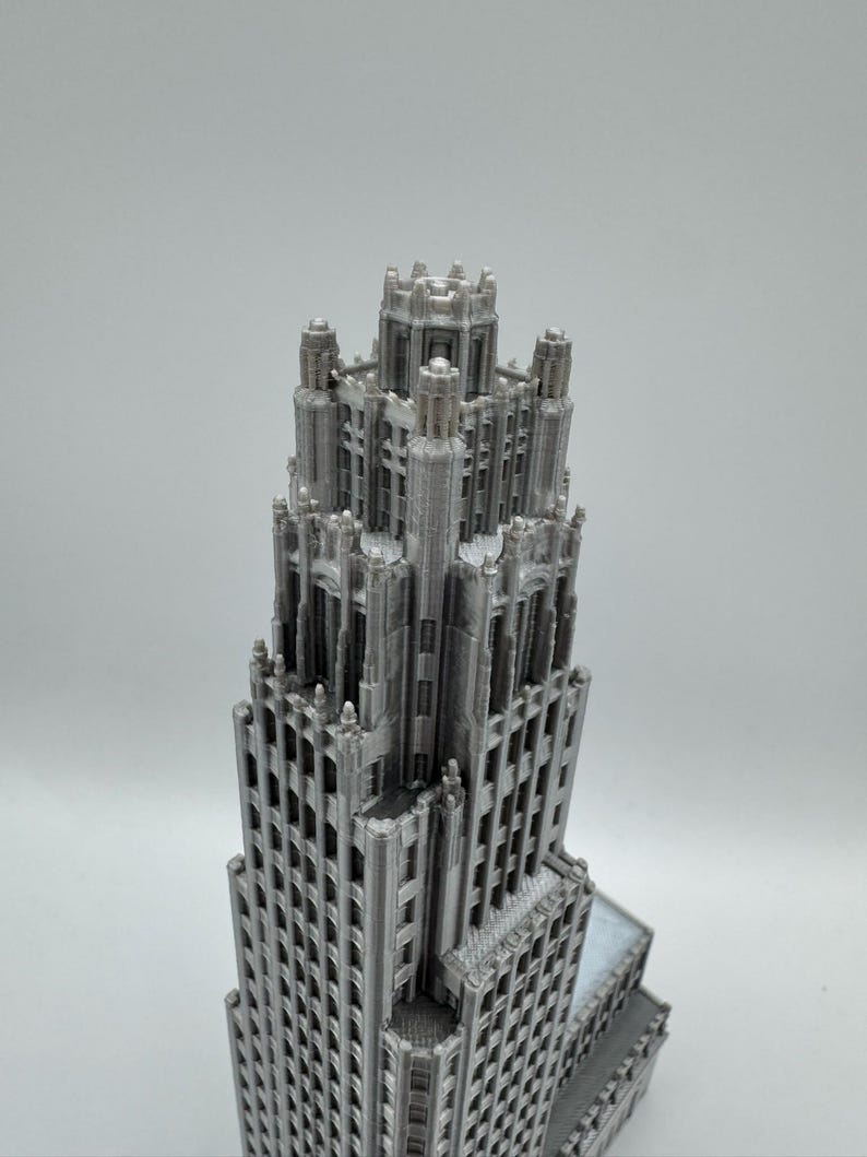 May include: A detailed, gray model of a skyscraper, showcasing intricate architectural details. The building features numerous windows and a tiered design, with a complex crown. The model is likely 3D-printed, with visible layer lines. A miniature replica of a famous building.