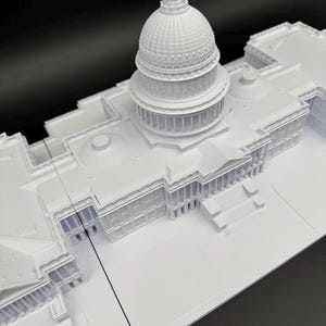 K&ouml;nnte beinhalten: Ein wei&szlig;es Architekturmodell des US-Kapitolgeb&auml;udes. Das Modell zeigt detaillierte Merkmale der Kuppel, der S&auml;ulen und der Gesamtstruktur. Das Modell befindet sich vor einem schwarzen Hintergrund.