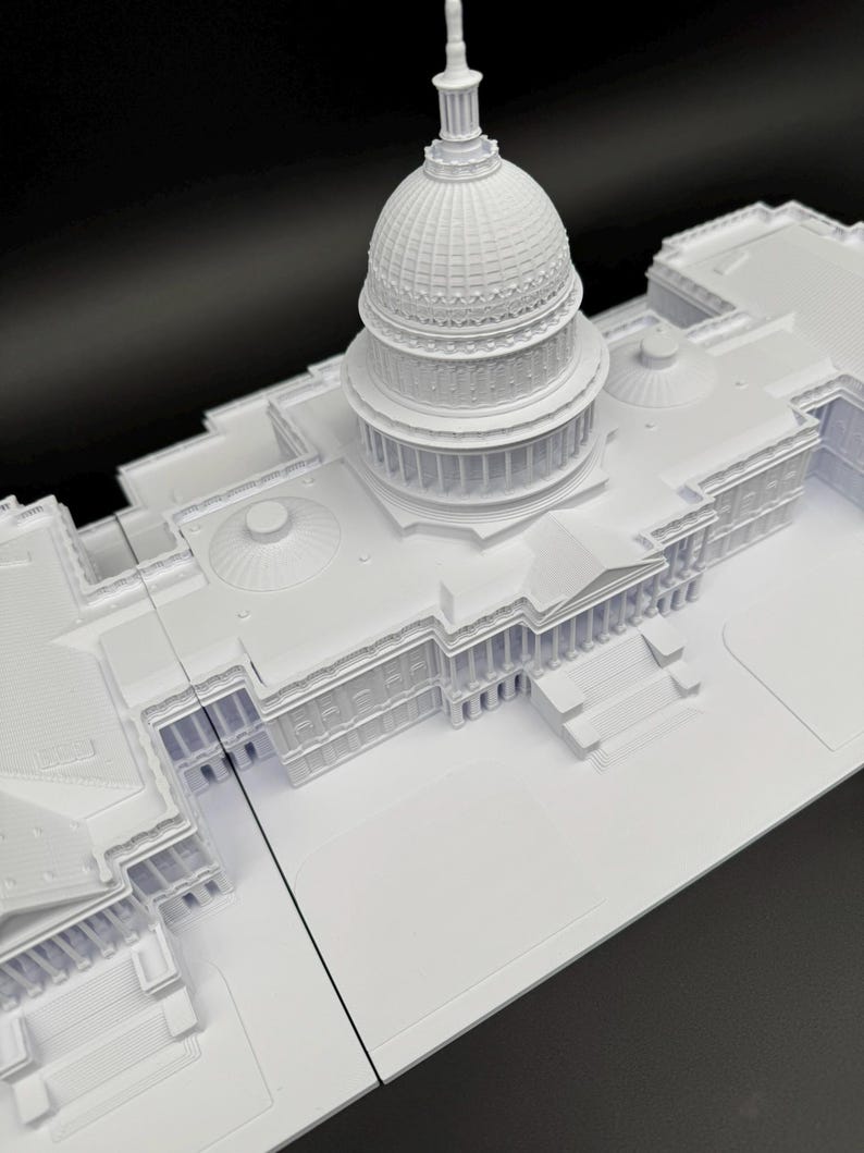 K&ouml;nnte beinhalten: Ein wei&szlig;es Architekturmodell des US-Kapitolgeb&auml;udes. Das Modell zeigt detaillierte Merkmale der Kuppel, der S&auml;ulen und der Gesamtstruktur. Das Modell befindet sich vor einem schwarzen Hintergrund.