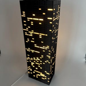 Peut inclure: Lampe noire rectangulaire en forme de gratte-ciel. Elle pr&eacute;sente de nombreuses petites fen&ecirc;tres rectangulaires &eacute;clair&eacute;es qui &eacute;mettent une lumi&egrave;re jaune chaude. La lampe est branch&eacute;e &agrave; un cordon blanc.