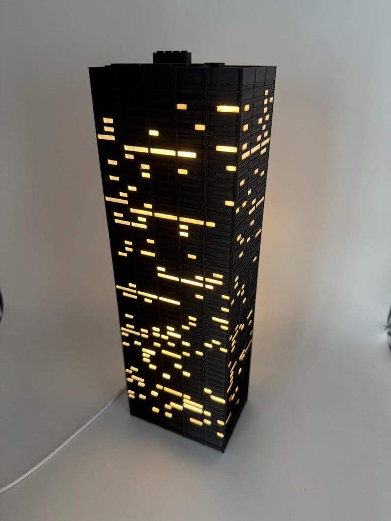 Peut inclure: Lampe noire rectangulaire en forme de gratte-ciel. Elle pr&eacute;sente de nombreuses petites fen&ecirc;tres rectangulaires &eacute;clair&eacute;es qui &eacute;mettent une lumi&egrave;re jaune chaude. La lampe est branch&eacute;e &agrave; un cordon blanc.
