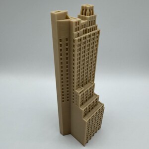 May include: A beige architectural model of a skyscraper. The building features a tall, rectangular tower with numerous windows, and a stepped design towards the base. The top of the building has a decorative crown.