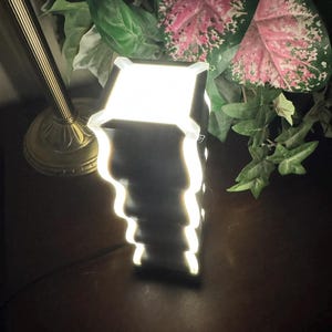 K&ouml;nnte beinhalten: Eine schwarz-wei&szlig;e Lampe mit einem einzigartigen, geschichteten Design. Die Lampe strahlt ein helles, warmes Licht aus und erzeugt einen sanften Schein. Die Lampe steht auf einer dunklen Holzoberfl&auml;che, im Hintergrund gr&uuml;ne und rosa Bl&auml;tter.