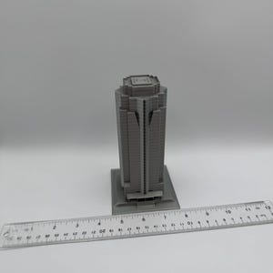 May include: A gray, miniature skyscraper model with a rectangular base. The building has a detailed facade with vertical lines and a flat roof. A ruler is placed next to the model, showing measurements in inches.