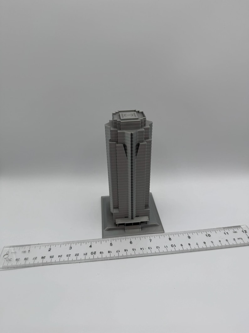 May include: A gray, miniature skyscraper model with a rectangular base. The building has a detailed facade with vertical lines and a flat roof. A ruler is placed next to the model, showing measurements in inches.