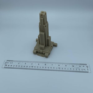 May include: A 3D printed model of a tall building with a pointed roof and many windows. The model is beige and is sitting on a white surface.