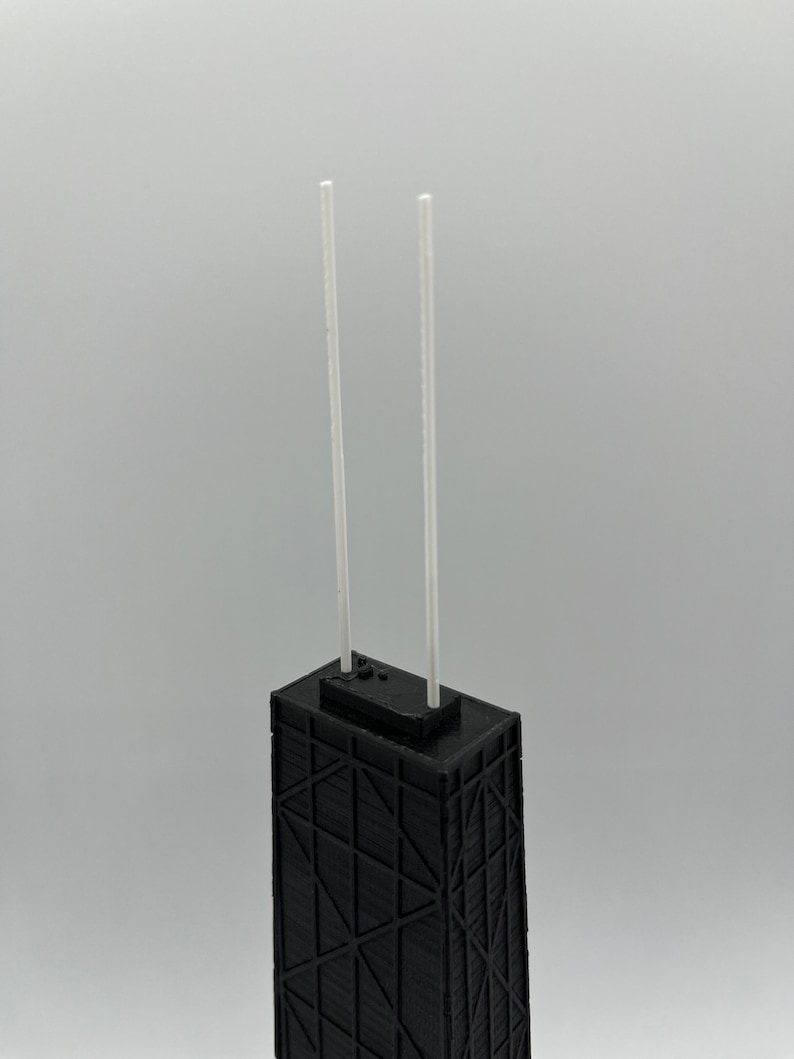 May include: A black architectural model of a skyscraper with two white antennae. The building has a geometric facade design and a flat roof. The model is set against a plain, light gray background.