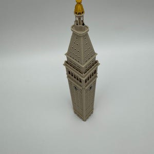 May include: A miniature model of the Metropolitan Life Insurance Company Tower, featuring a light gray structure with a square base, a tapered clock tower section, and a prominent gold-colored spire at the very top of the architectural replica.