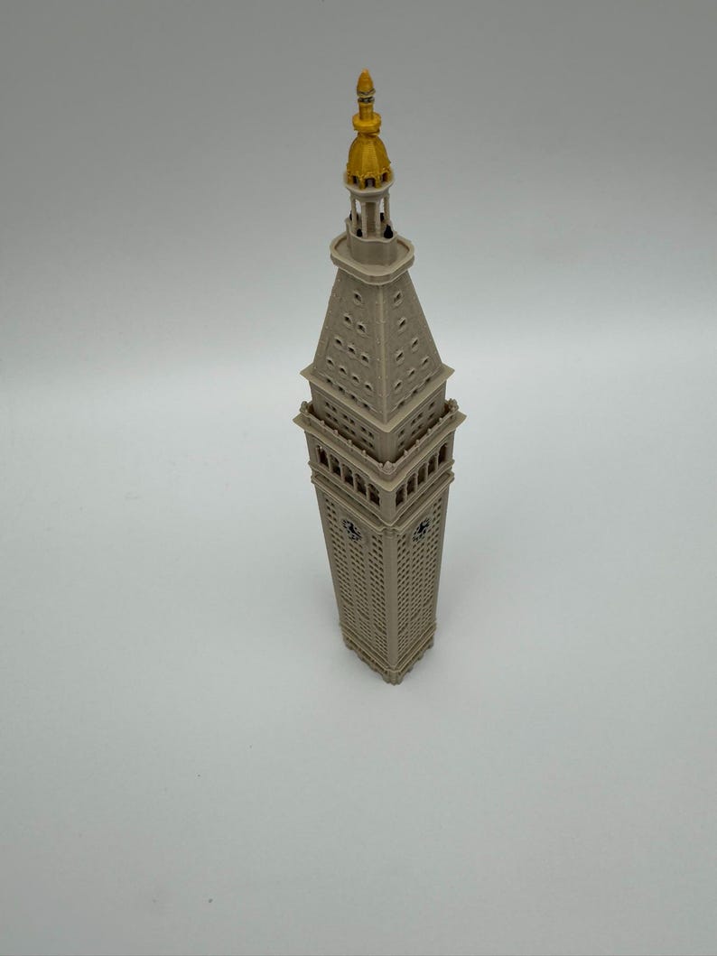 May include: A miniature model of the Metropolitan Life Insurance Company Tower, featuring a light gray structure with a square base, a tapered clock tower section, and a prominent gold-colored spire at the very top of the architectural replica.