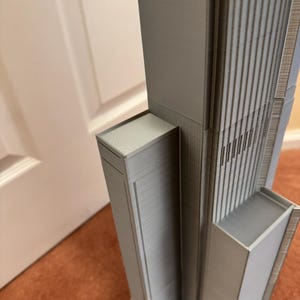 K&ouml;nnte beinhalten: Graues Architekturmodell eines Wolkenkratzers mit detaillierten vertikalen Linien und rechteckigen Strukturen. Das Modell ist hellgrau und scheint 3D-gedruckt zu sein. Das Design umfasst mehrere Ebenen und eine strukturierte Oberfl&auml;che.