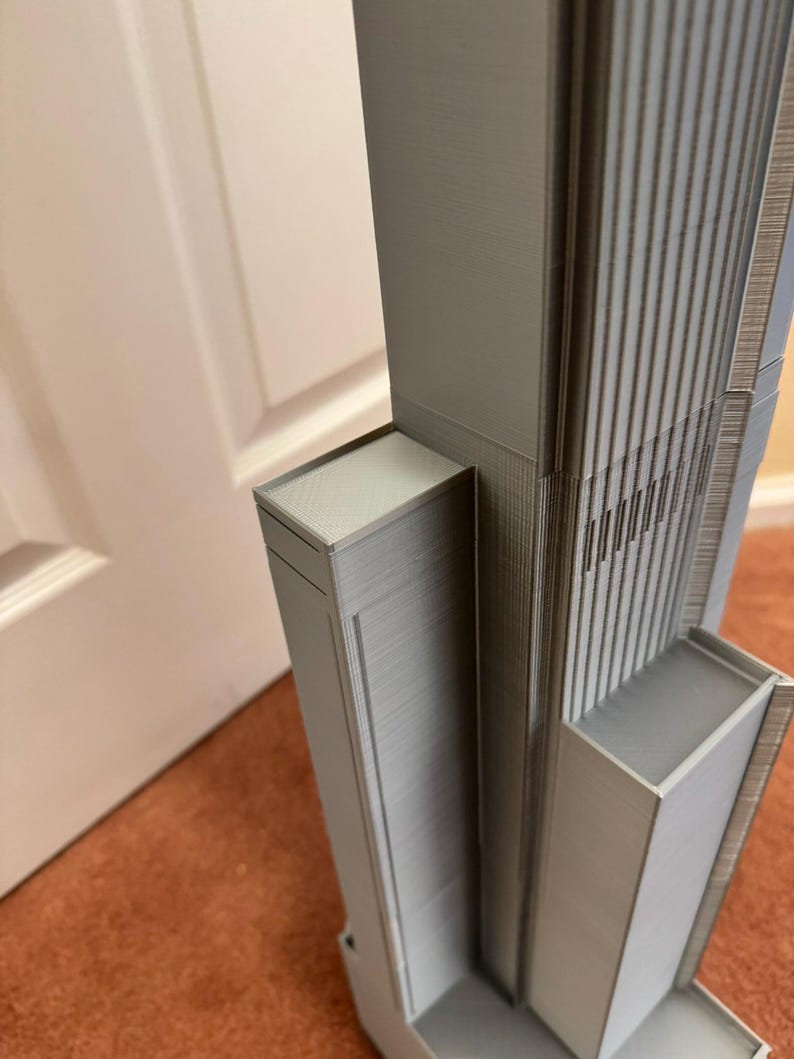 K&ouml;nnte beinhalten: Graues Architekturmodell eines Wolkenkratzers mit detaillierten vertikalen Linien und rechteckigen Strukturen. Das Modell ist hellgrau und scheint 3D-gedruckt zu sein. Das Design umfasst mehrere Ebenen und eine strukturierte Oberfl&auml;che.