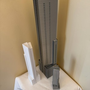 May include: Three miniature skyscraper models in gray and white are displayed on a white surface. A ruler is placed in front of the models. The largest model is gray and has a tall, rectangular shape with multiple windows. The other two models are smaller, one white and one gray.