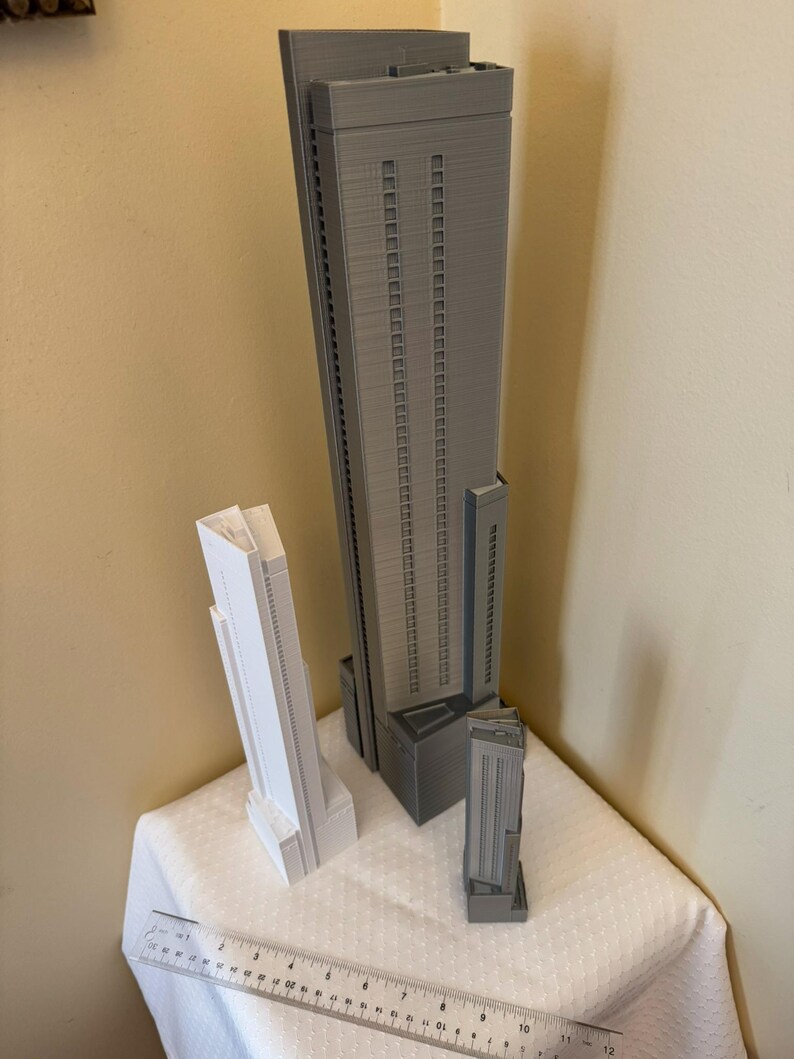 May include: Three miniature skyscraper models in gray and white are displayed on a white surface. A ruler is placed in front of the models. The largest model is gray and has a tall, rectangular shape with multiple windows. The other two models are smaller, one white and one gray.