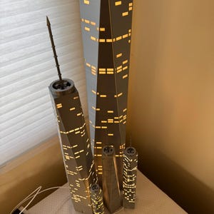 May include: A collection of gray and white skyscraper lamps with illuminated windows. The lamps vary in size, with the tallest one measuring approximately 24 inches. The lamps are arranged on a white surface, with a ruler and power cord visible.
