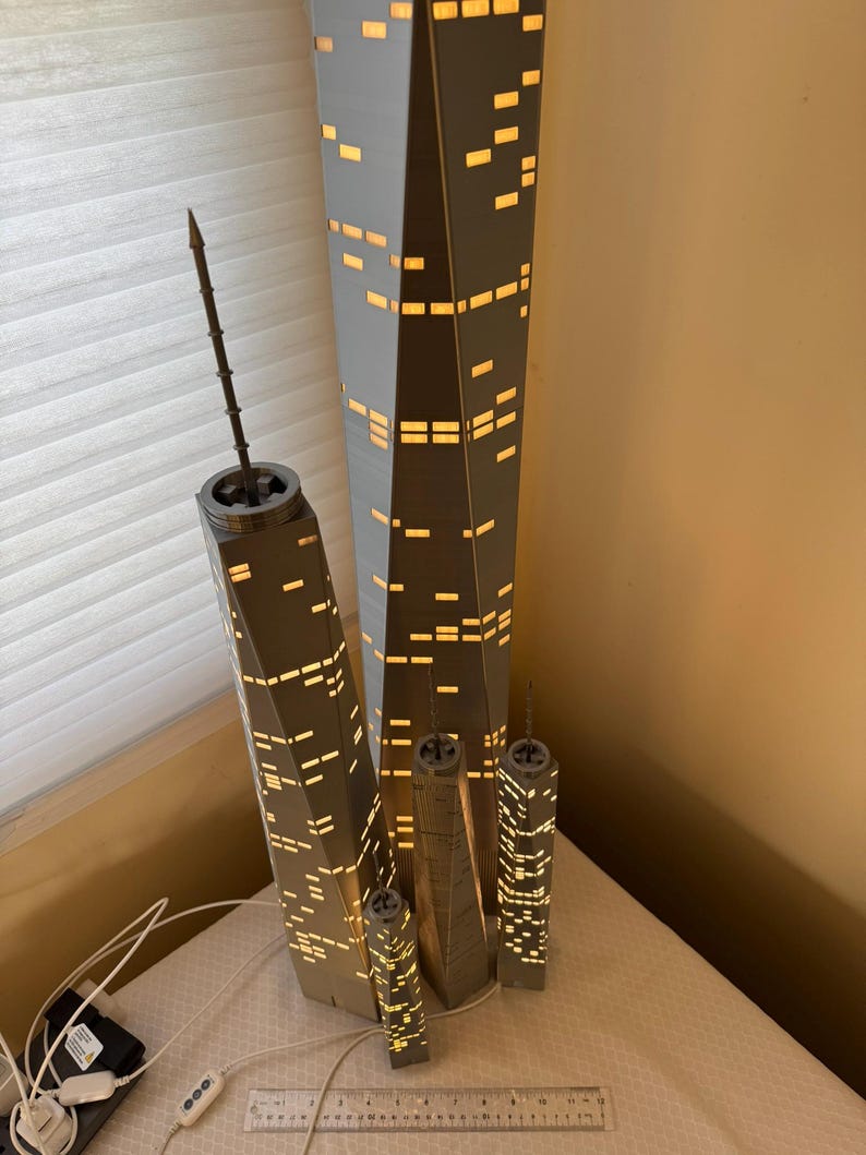 May include: A collection of gray and white skyscraper lamps with illuminated windows. The lamps vary in size, with the tallest one measuring approximately 24 inches. The lamps are arranged on a white surface, with a ruler and power cord visible.