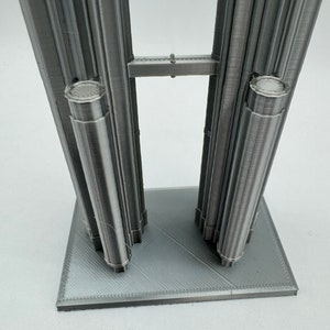 May include: A 3D printed model of two tall buildings connected by a bridge. The buildings are made of a silver-colored material and have a textured surface. The model is sitting on a gray base.