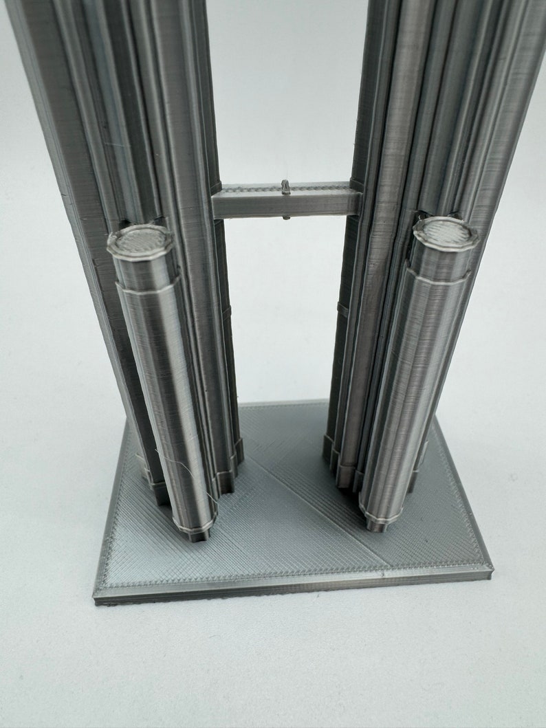 May include: A 3D printed model of two tall buildings connected by a bridge. The buildings are made of a silver-colored material and have a textured surface. The model is sitting on a gray base.