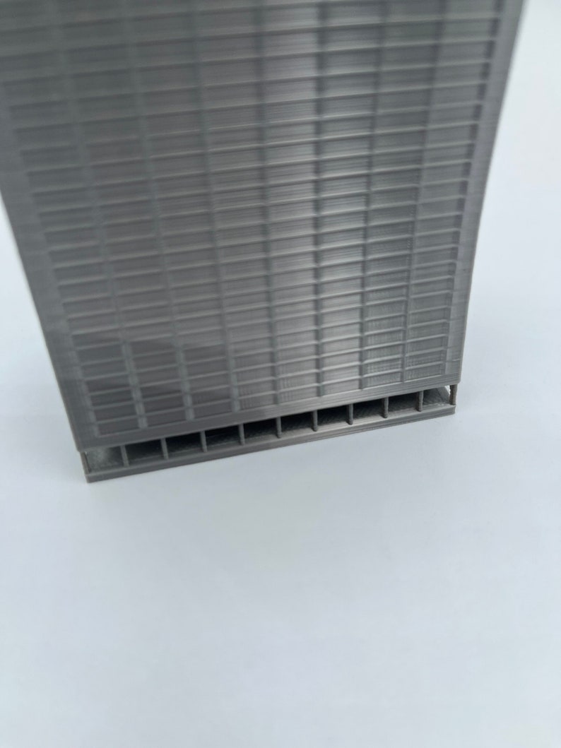 May include: A gray 3D printed model of a skyscraper with a grid pattern on the facade. The model has a base with a series of small openings.